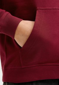 Sudadera burdeos con bolsillo canguro, puños y dobladillo acanalados. Textura suave con detalles de costura a lo largo de las costuras.