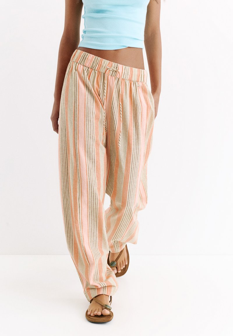 Femme portant un pantalon ample rayé pêche et beige, un top court bleu clair et des sandales marron avec un détail en pierre verte, marchant sur un sol blanc.