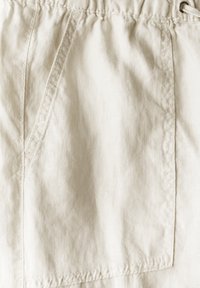 Ljust beige tygshorts med en sidficka, elastisk midja med dragsko och synliga sömdetaljer. Strukturerat linne-material.