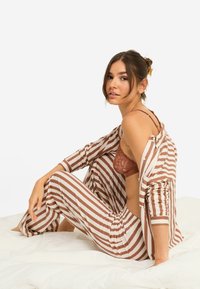 Σετ σαλονιού με ρίγες σε εκρού και καφέ, που περιλαμβάνει πουκάμισο με κουμπιά και παντελόνια φαρδιάς γραμμής. Υφή δαντέλας σε σκουριά εσώρουχο από κάτω, τοποθετημένο σε λευκό κρεβάτι.