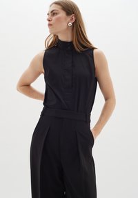 Blusa nera senz maniche con colletto alto e chiusura a bottoni, abbinata a pantaloni neri a vita alta e plissettati. Tessuto morbido.