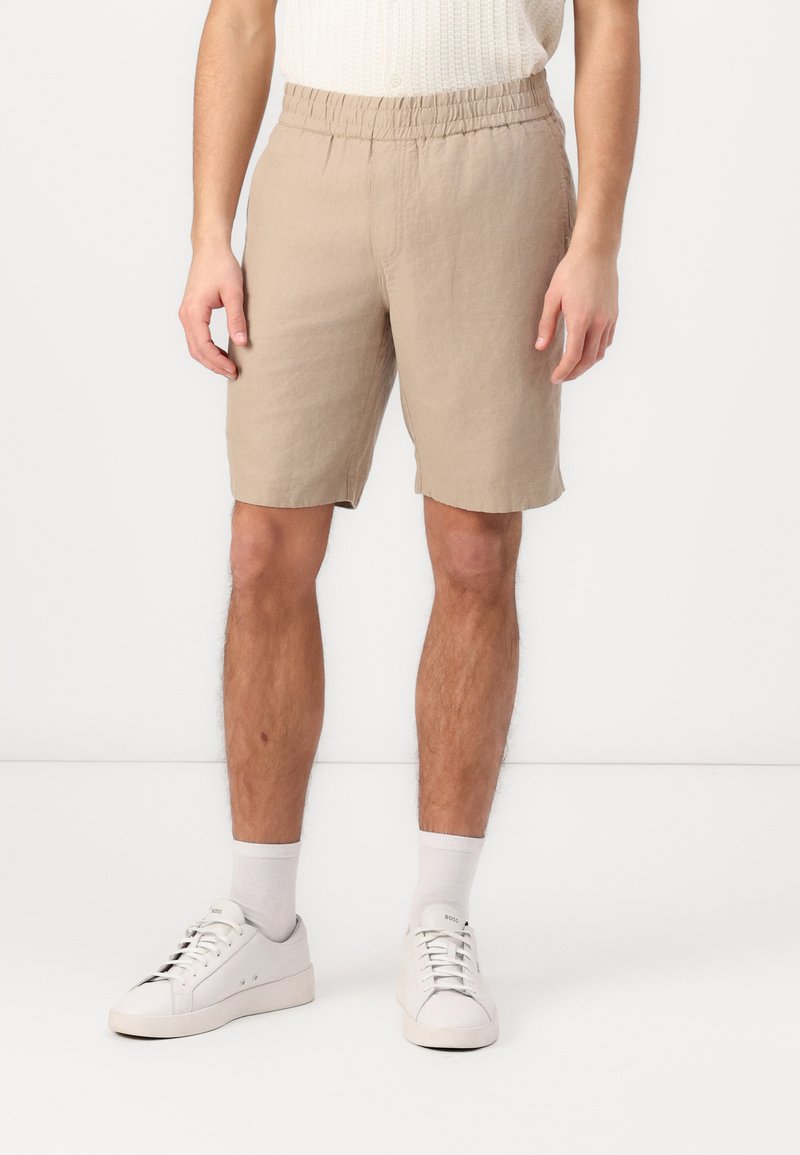 Pantalones cortos de lino beige con cintura elástica, que caen por encima de la rodilla. Combinados con zapatillas blancas y calcetines gris claro. Diseño simple y casual.
