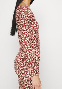 Vestido floral con una silueta ajustada, mangas largas y detalles fruncidos. La tela tiene un fondo rosa con patrones florales en rojo y verde.