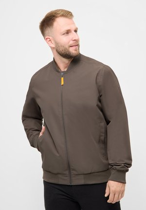 Mand iført brun bomberjakke med lynlås og ribstrikkede manchetter, ser mod højre med hånden i lommen mod en ensfarvet hvid baggrund.