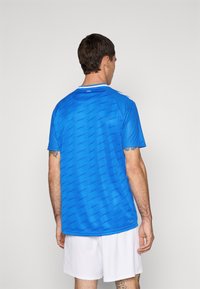 Man met zijn rug naar de kijker, gekleed in een blauwe sportoverhemd met een patroon en "1965" op de kraag, en witte shorts, tegen een effen lichte achtergrond.