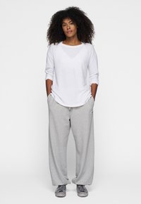 Camicia bianca a maniche lunghe con scollo rotondo, abbinata a pantaloni sportivi grigi larghi con cordoncino e una patch logo laterale. Sneakers grigie.