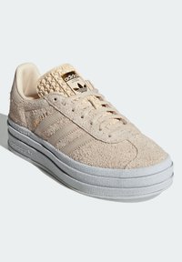 Beige suede sneaker with three horizontal stripes, textured tongue detail, gold logo on the tongue, and a gray rubber sole.