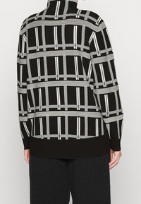 Pull en noir et gris à motifs, avec un col haut, des manches longues et des rayures géométriques. Texture tricotée douce avec une coupe ample.