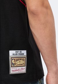 Μαύρη φανέλα μπάσκετ με "1997-98 Allen Iverson" και το έμβλημα NBA Hardwood Classics Mitchell & Ness κοντά στο κάτω άκρο δίπλα στο μπράτσο ενός ατόμου.