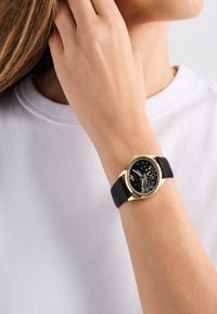 Schwarze Lederarmbanduhr mit einem Gehäuse aus goldfarbener Legierung, schwarzes Zifferblatt mit floralen Mustern und metallischen Akzenten, getragen an einem Handgelenk mit einem weißen Hemd.