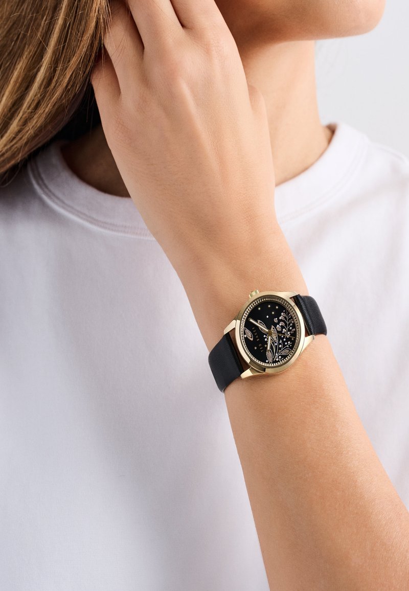 Schwarze Lederarmbanduhr mit einem Gehäuse aus goldfarbener Legierung, schwarzes Zifferblatt mit floralen Mustern und metallischen Akzenten, getragen an einem Handgelenk mit einem weißen Hemd.