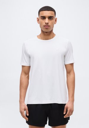 Jauns vīrietis stāv, skatoties tieši priekšā, valkājot vienkāršu baltu īsrokas kreklu un melnas šortus gaiši pelēka fona priekšā.