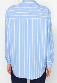 Ljusblå långärmad skjorta med vita vertikala ränder, en knapp-ner krage och rundad nederkant. Mjuk, slät tyg.