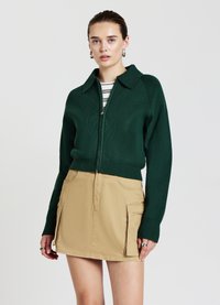 Maglione verde lavorato a maglia con zip e colletto, abbinato a una mini gonna beige con tasche laterali e una trama liscia.