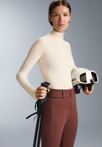 Kremasti rebrasti pleteni pulover s visokim ovratnikom, prilagođenog kroja, u kombinaciji s hlačama za skijanje u visokim strukom u crvenkasto-smeđoj boji; drži crne skijaške štapove i bijelu kacigu.