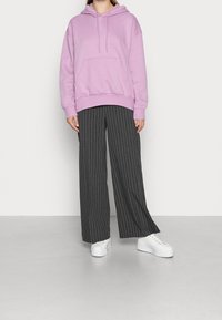 Sudadera de color púrpura claro con bolsillo frontal, combinada con pantalones anchos de rayas grises y zapatillas de plataforma blancas. Textura suave, corte relajado.