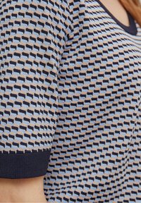 Detailní záběr na vzorovaný svetr s geometrickým designem v béžové, modré a námořnické barvě, zvýrazňující texturu a výstřih.