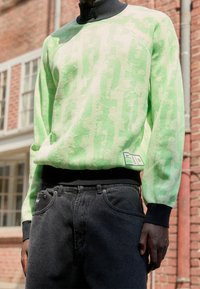 Maglione verde e crema con un motivo texturizzato, colletto alto e polsini in nero a costine. Abbinato a jeans neri con una vestibilità classica.