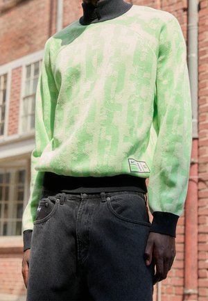 Suéter verde y crema con un patrón texturizado, cuello alto y puños de canalé negros. Combinado con unos jeans negros de corte clásico.