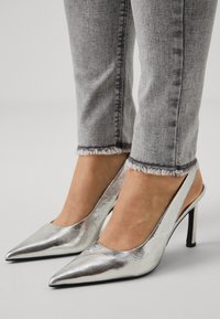 Zapatos de tacón alto plateados con tiras en el talón, que presentan una punta afilada, textura metálica y tacón de aguja, combinados con jeans gris deshilachados.