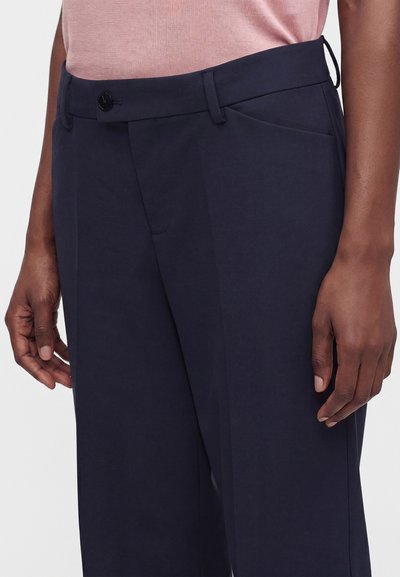 Pantalon bleu marine foncé avec passants de ceinture, fermeture à boutons noirs, poches latérales, porté par une personne à la peau brune et un haut rose.