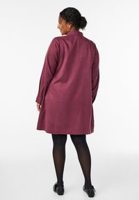 Kihara naise, kellel on kuklas lokkis juuksed, kannab pika varrukaga põlvepikkust burgundia värvi kleiti, musti retuuse ja musti tasa kingi, seistes seljaga. 