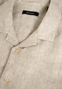 Camicia beige in lino a maniche corte con colletto notch, dotata di chiusura a bottoni e un'etichetta nera con testo bianco attaccata al collo.