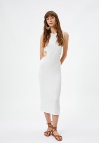 Witte, geribbelde gebreide jurk met een mouwloos ontwerp, uitsparingen aan de zijkant en een lengte tot de knie. Model gecombineerd met bruine sandalen met bandjes en grote oorbellen.
