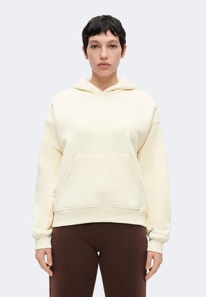 Personne aux cheveux courts et foncés portant un sweat à capuche couleur crème et un pantalon marron foncé, debout devant un fond clair uni.