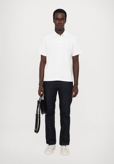 Polo shirt blanc à manches courtes avec trois boutons, associé à un jean bleu foncé et des baskets. Tient un sac à main noir avec une bandoulière.
