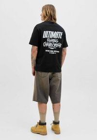Persona con tatuajes lleva una camiseta negra de "Ultimate Fighting Championship", pantalones cortos grises, calcetines beige y botas de trabajo marrones, de espaldas.