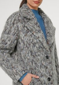 Grau, strukturierten Mantel mit mehrfarbigen Fasern, großen Revers, zwei Knöpfen und seitlichen Taschen. Kombiniert mit einem blauen Pullover darunter.