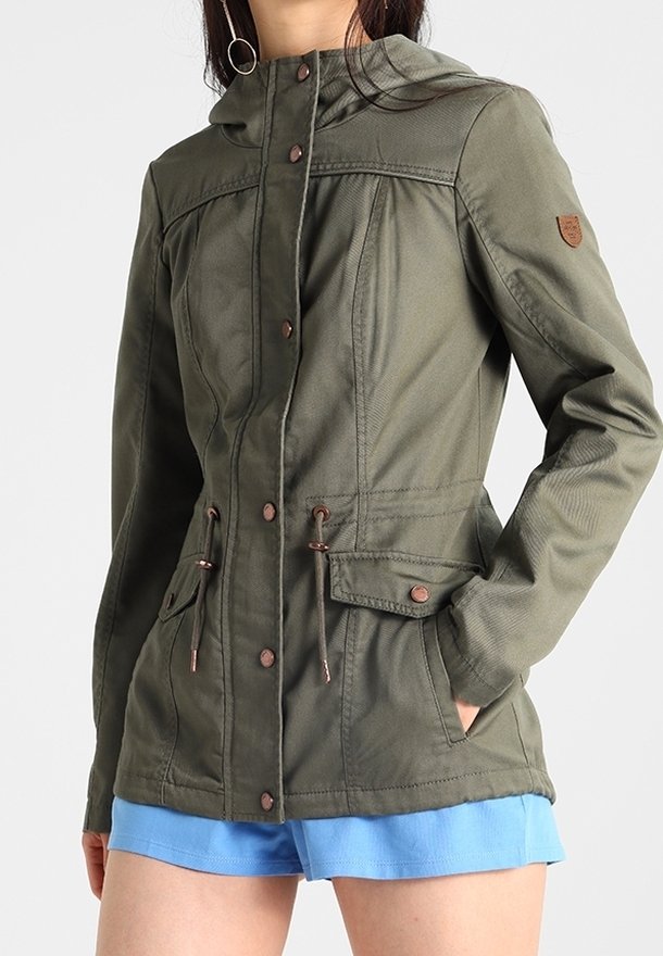 Femme portant une veste à capuche vert olive avec boutons à l'avant, taille réglable par cordon, poches à rabat, et un short bleu sur un fond uni.