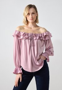 Bluza bez naramenica od mekog ružičastog materijala s volanima na dekolteu i dugim rukavima; sadrži vezicu i naborane manžete.