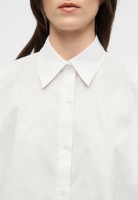 Femme portant une chemise blanche boutonnée avec un large col pointu et des motifs subtils en forme de cœur sur le tissu.