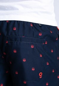 Pantaloni da bagno navy con motivo a sole e onde rosse, vita elastica, tasca posteriore e un piccolo logo. Presenta una grommet decorativa.