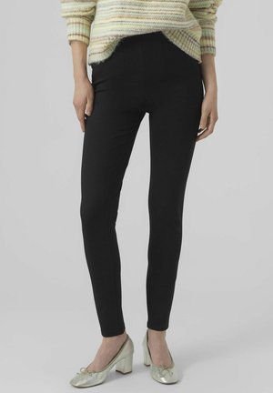 Pantaloni stretch neri con una vestibilità slim, caratterizzati da una vita alta e linee pulite, abbinati a scarpe con tacco a blocco in argento lucido.