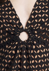Haut noir avec un design texturé et découpé, présentant un rassemblement circulaire central. Le tissu semble doux et légèrement structuré.
