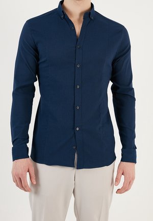 Mann trägt ein eng anliegendes dunkelblaues Hemd mit langen Ärmeln und hellbeige Hose vor einem einfarbigen Hintergrund.