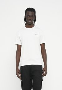 Λευκό βαμβακερό t-shirt με κοντά μανίκια, στρογγυλή λαιμόκοψη και ένα μικρό μαύρο λογότυπο στο στήθος. Συνδυασμένο με μαύρα cargo σορτς.