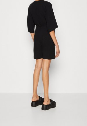 Salopette nera con maniche corte e vita cinchata, caratterizzata da una texture liscia e una vestibilità comoda. Abbinata a sandali platform neri chunky.