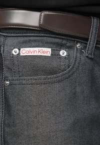 Melni džinsa bikses ar teksturētu apdari, uz kurām ir sarkani un balti Calvin Klein uzraksti, sudraba kniedes un brūns ādas josta.