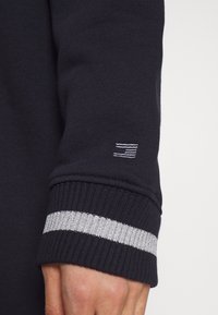 Marinblå sweatshirtmanschett med en silverrand och en subtil logotyp broderad i vit tråd. Slät stickad yta med ribbade kanter.