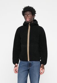 Svart fleecejakke med hette, med en fargerik stripete glidelås. Stoffet er mykt og teksturert, med ribbede mansjetter og fald.
