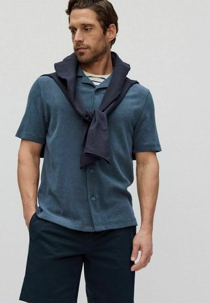 Mannenshirt met korte mouwen in teal met een textuurafwerking, voorzien van een knoopsluiting en een marineblauwe sweater die rond de schouders is geknoopt. Donkere shorts.