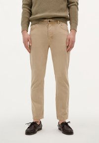 Pantalones de mezclilla de algodón beige con un corte ajustado, diseño de cinco bolsillos y cierre de botón. Llevados con zapatos oscuros de cordones.