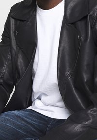 Giacca da motociclista in pelle nera con colletto appuntito, dettagli in argento e tasche con zip. Sotto si intravede una t-shirt bianca a girocollo.