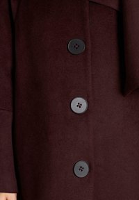 Bordeaux wollen jas met een gladde textuur, voorzien van vier zwarte knopen, elk met een contrasterend midden en vier gaten. Maatwerkontwerp met een revers.
