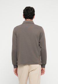 Mann med kort krøllete hår iført en brun langermet skjorte og beige bukser, stående med ryggen mot en ensfarget hvit bakgrunn.