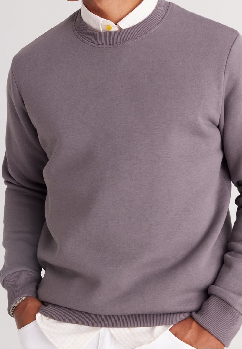 Homme portant un sweat-shirt gris ras du cou sur une chemise blanche boutonnée, les mains dans les poches d’un pantalon blanc devant un fond uni.